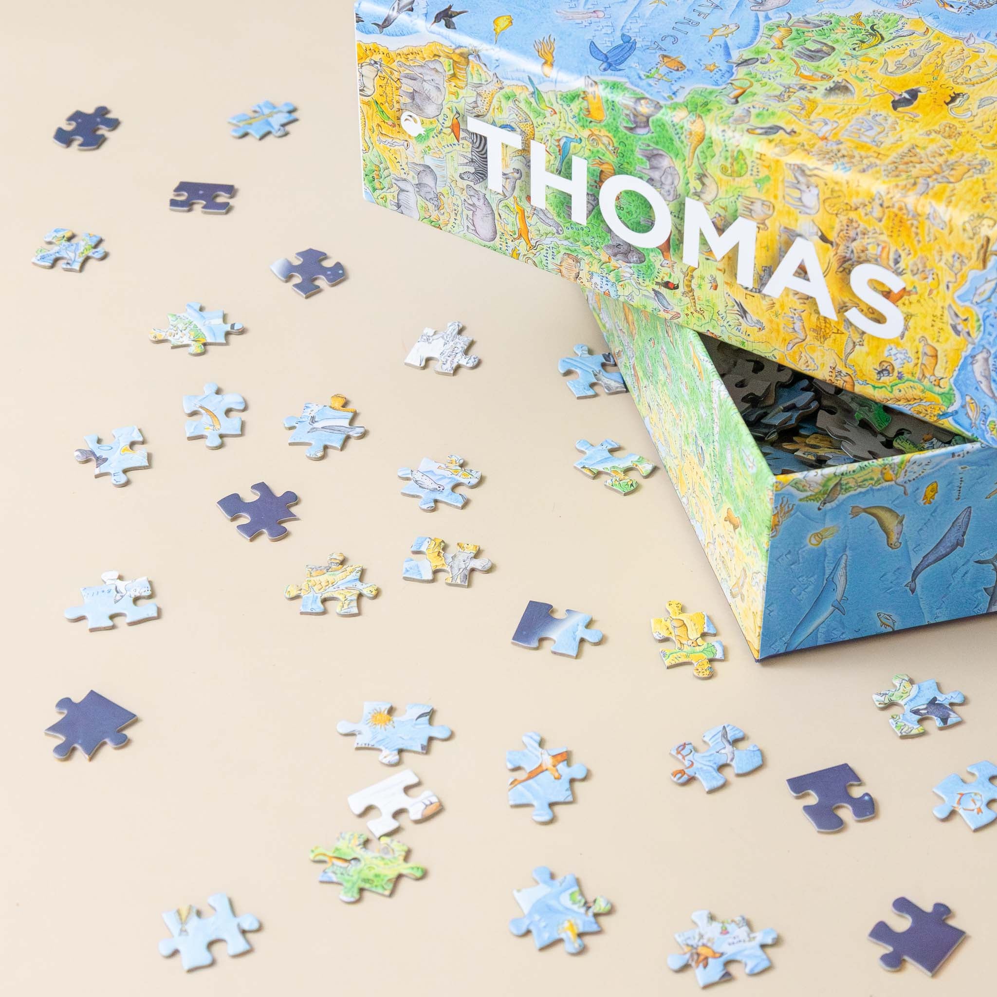 anton-thomas-1500-piece-wild-world-puzzle Jigsaw puzzle with pieces scattered on a beige surface, featuring a colorful box labeled 'THOMAS'.