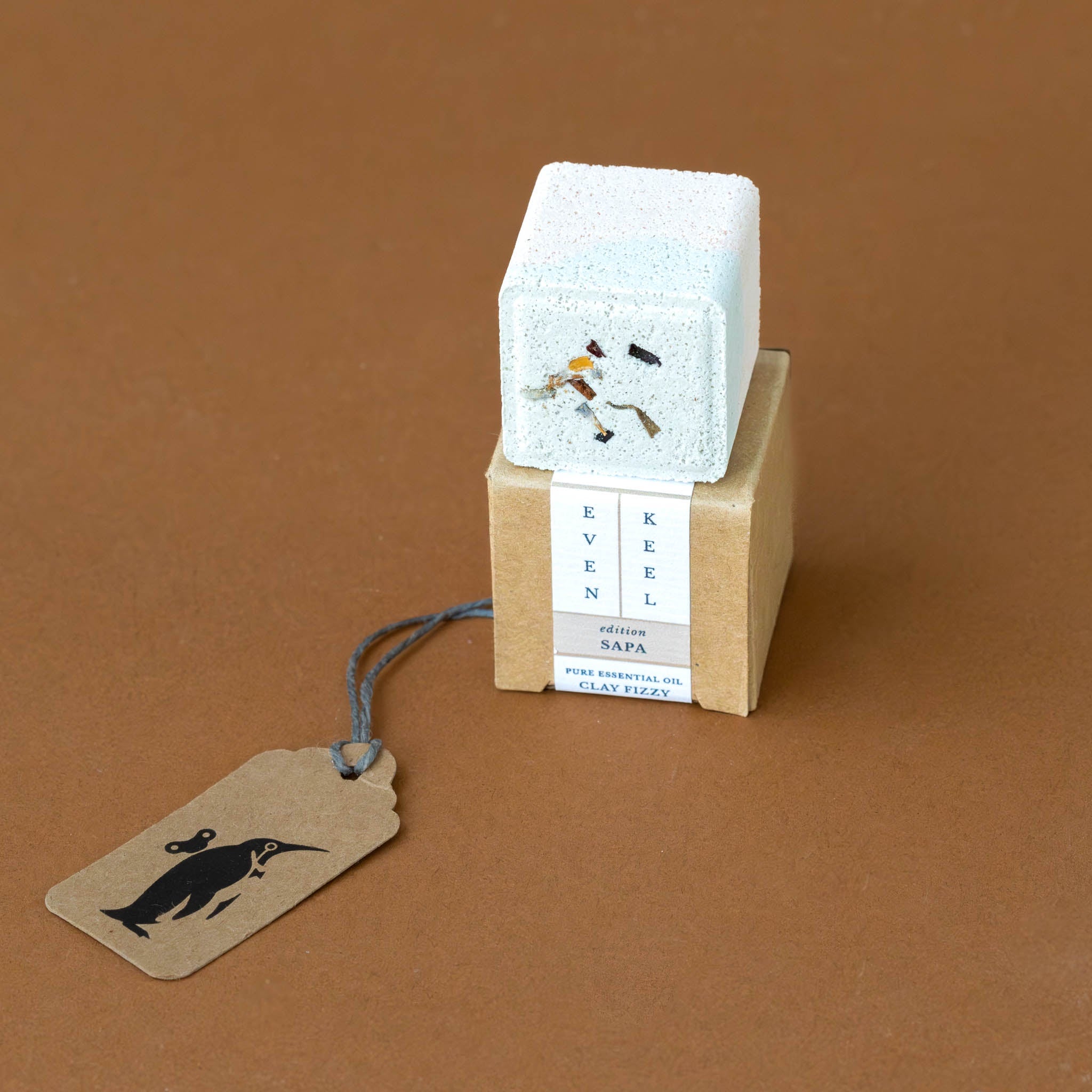 aromatherapy-bath-fizzy-cube-sapa A travel-sized aromatherapy bath fizzy cube with a white top, natural brown color, and a tag attached that reads 'Sapa'.