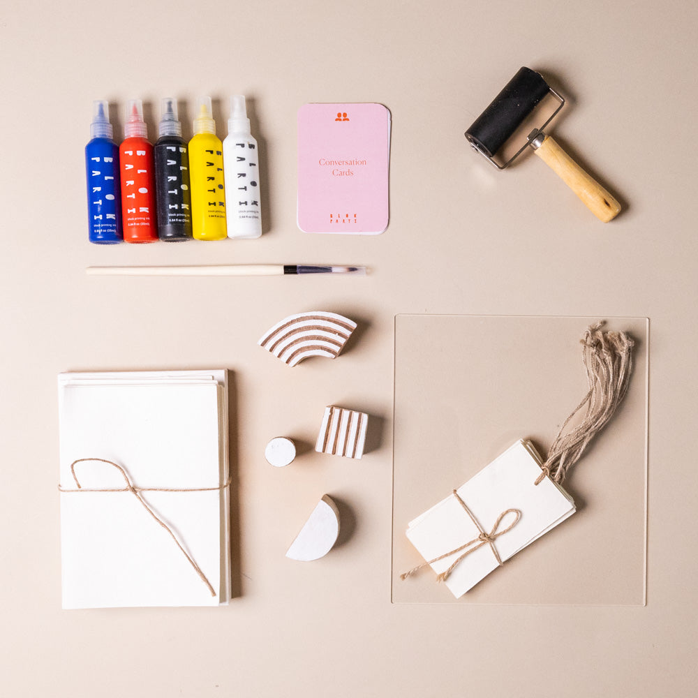 create-your-own-block-print-stationary-kit-abstract Collection of art supplies including paint bottles, a pink container, a brush, and other tools on a beige surface.