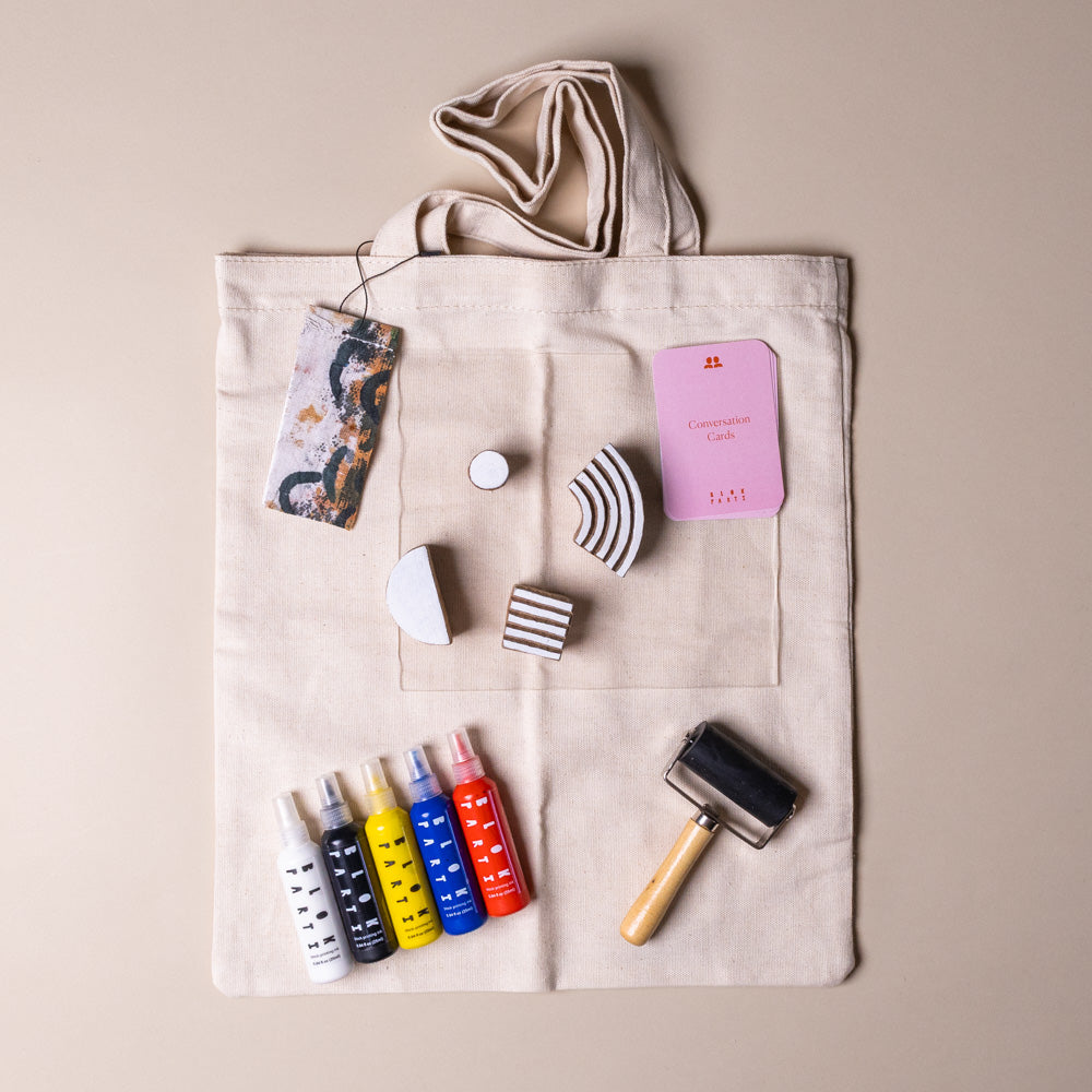 create-your-own-block-print-tote-kit-abstract Beige tote bag with art supplies including paint bottles, a brush, and small containers on a beige background