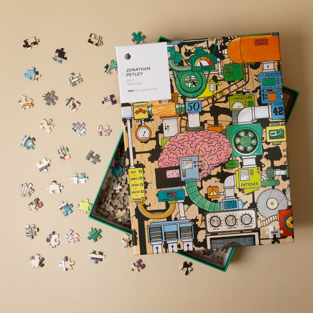 jonathan-petleys-self-machine-1000-piece-puzzle Puzzle box with colorful design and scattered puzzle pieces on a beige background