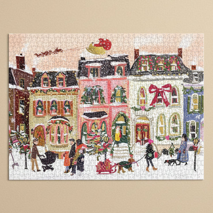 snowfall-on-main-street-1000-piece-puzzleChristmas-themed puzzle with snowy village scene and people, featuring a pink building with a large red bow.