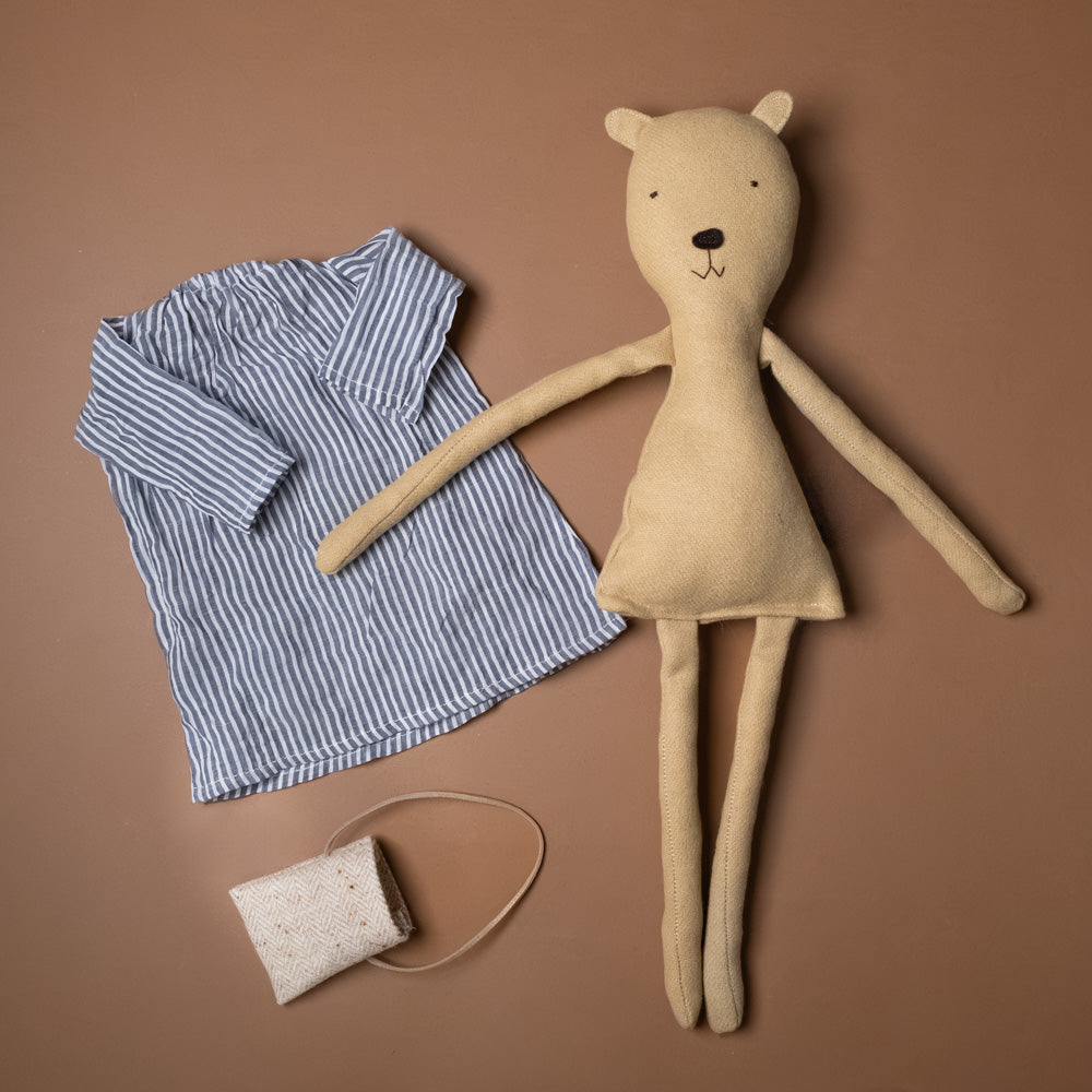 agatha-the-bear-chambray-stripe-day-dress-stuffed-animal Beige teddy bear, blue striped shirt, and beige bag on a brown background