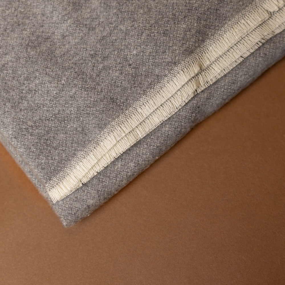 aspen-throw-ash Close-up of a gray fabric with a beige border on a brown background