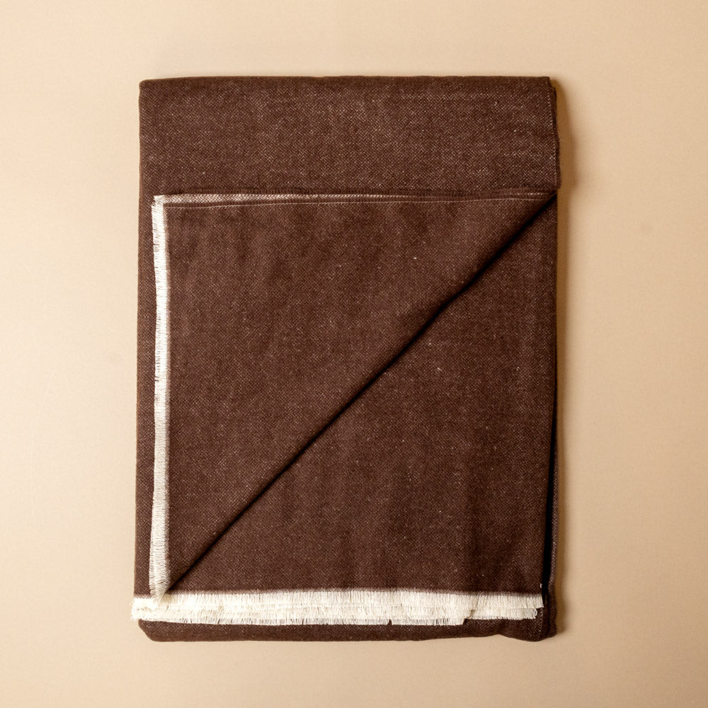 aspen-throw-chocolate Folded brown fabric with white trim on a beige background