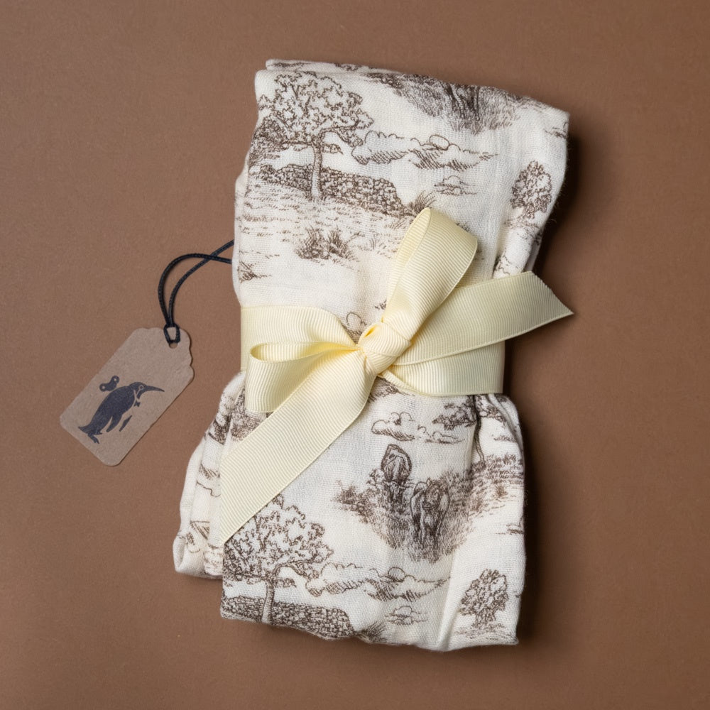 bamboo-burpie-set-meadow-toile Folded fabric with a scenic pattern tied with a yellow ribbon on a brown background