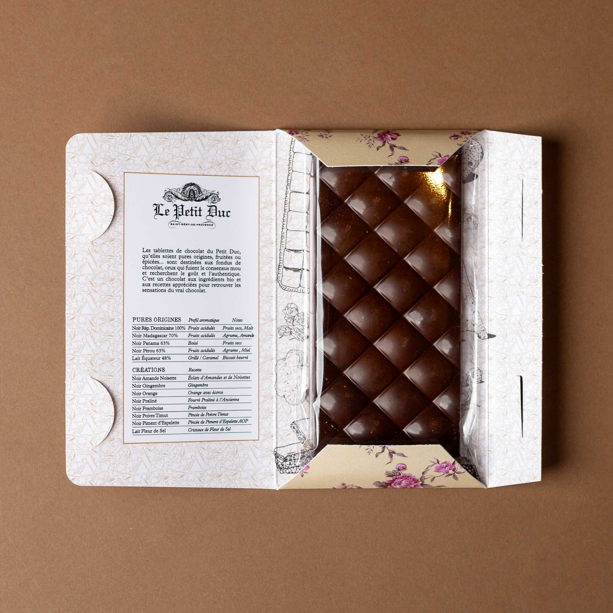 chocolat-noir-bar-almonds-and-hazelnuts Open package with a brown chocolate bar and decorative paper on a brown background