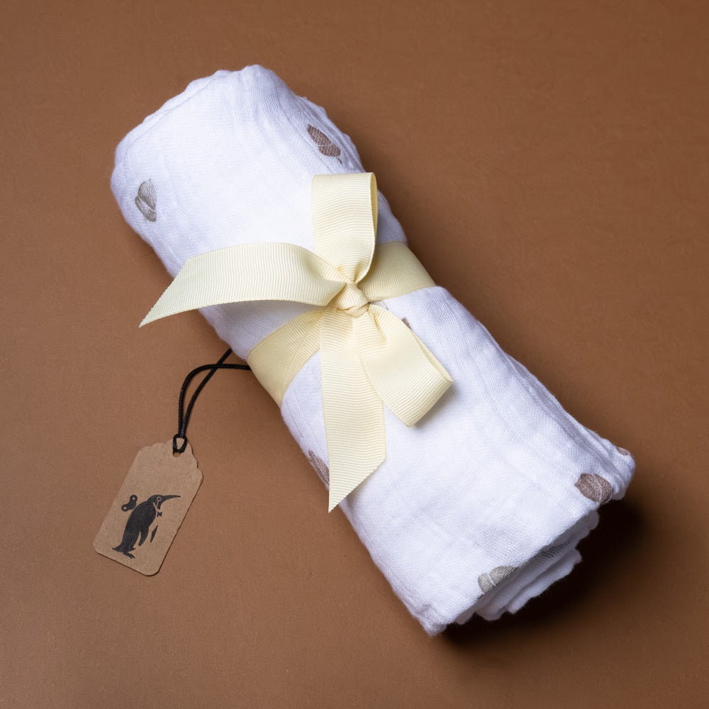 cotton-muslin-swaddle-acorns Rolled-up white swaddle with a yellow ribbon and tag on a brown background