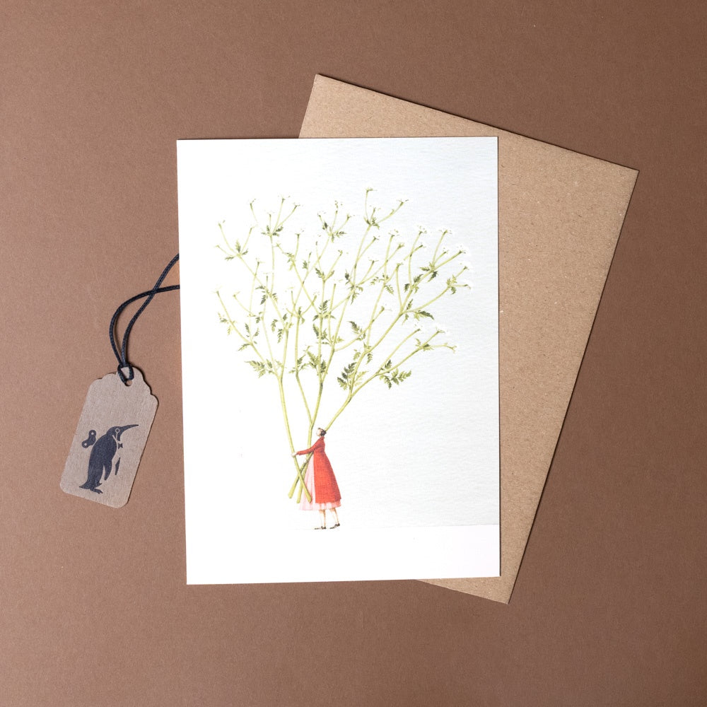 Greeting card with illustration of a person holding dried flowers, accompanied by a tag and brown envelope on a brown background.