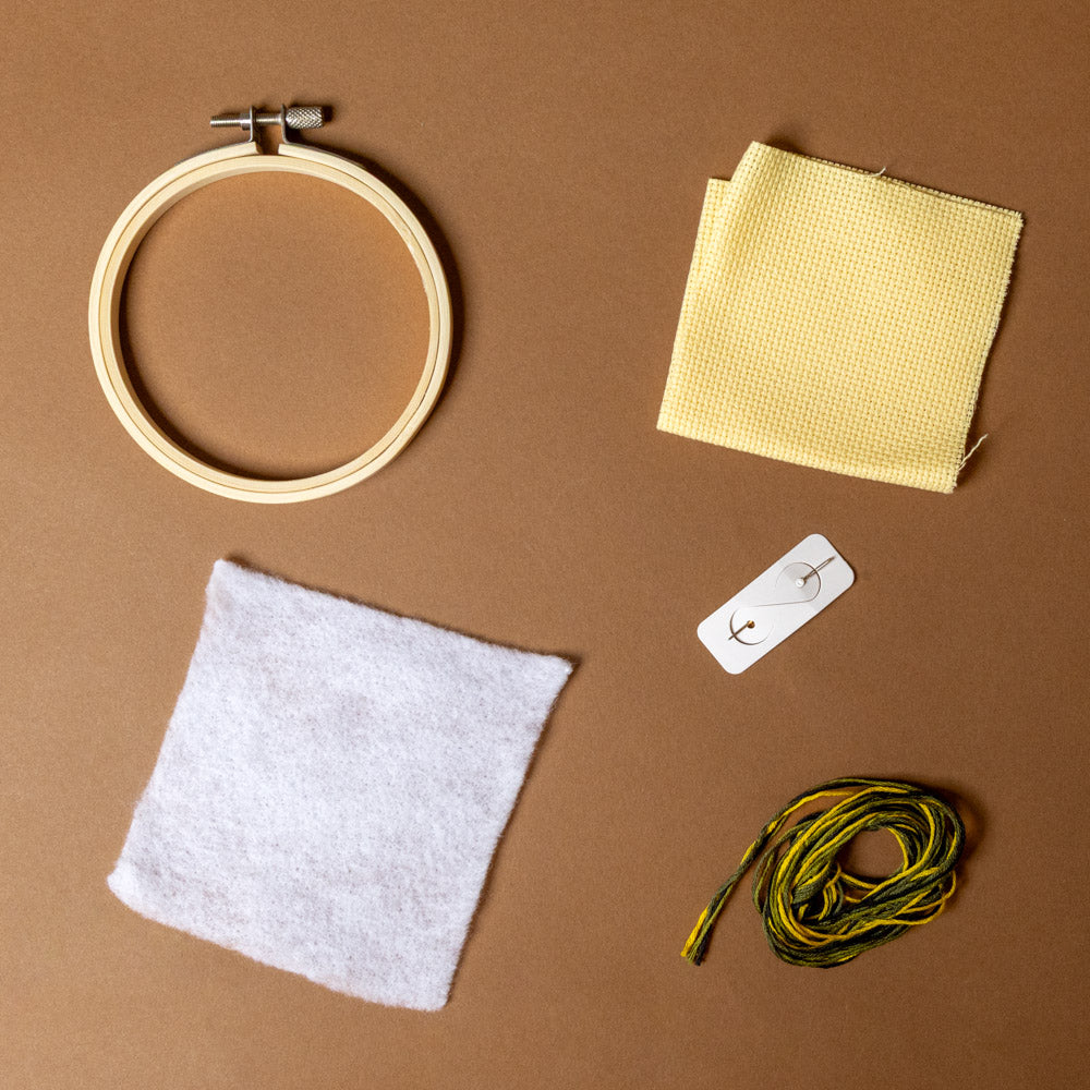 cross-stitch-kit--frog Embroidery hoop, fabric, floss, and needle on a brown background