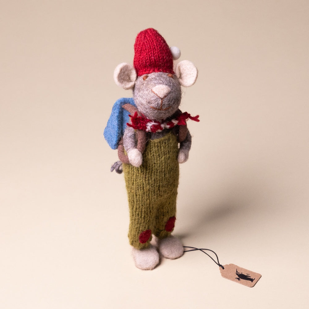 felted-grey-mouse-green-overalls-and-back-pack Knitted mouse toy with a red hat, blue backpack, and green outfit on a beige background