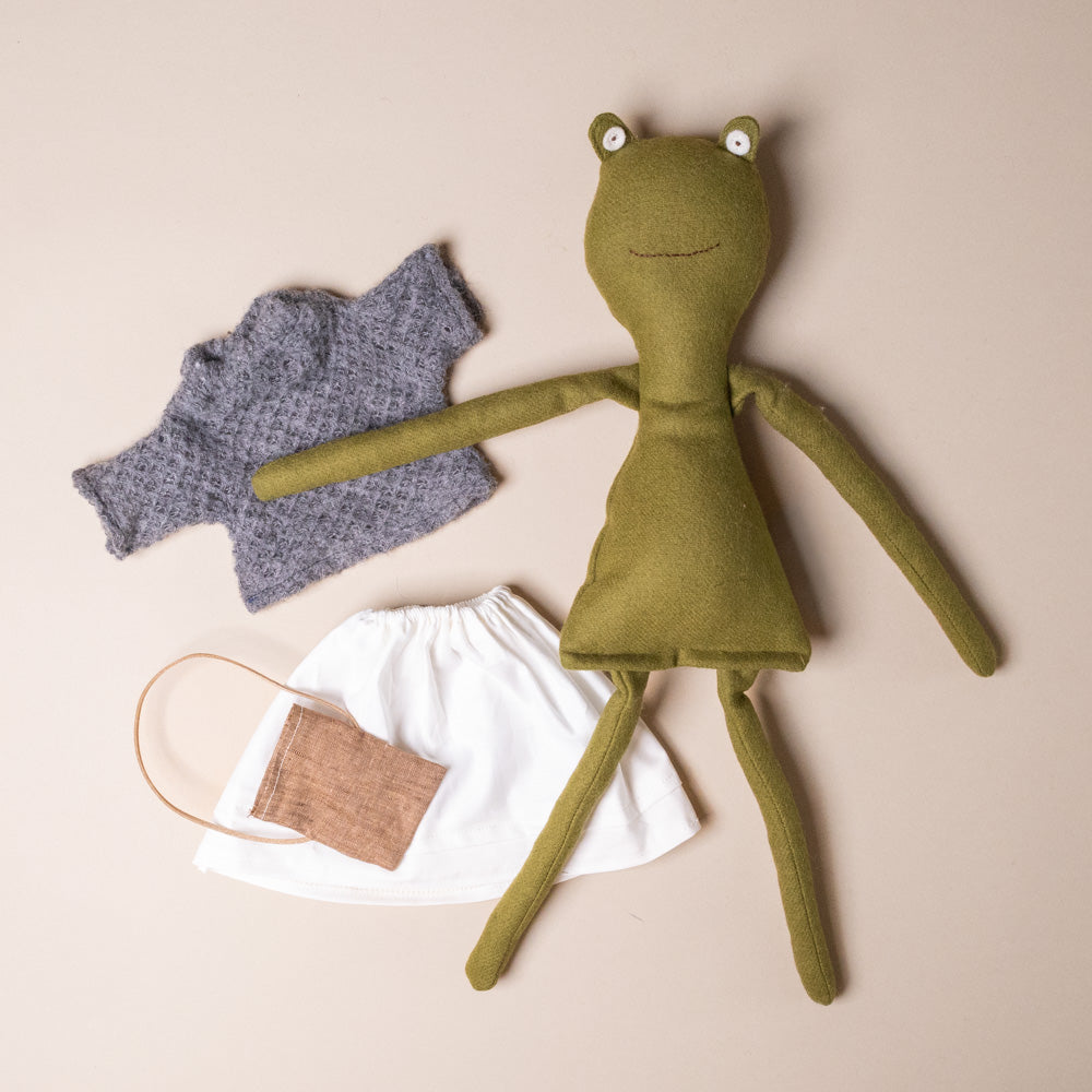 fern-the-frog-french-knit-sweater-and-morning-skirt-stuffed-animal-with-pink-satchel Green plush toy frog with a gray sweater and white drawstring bag on a beige background
