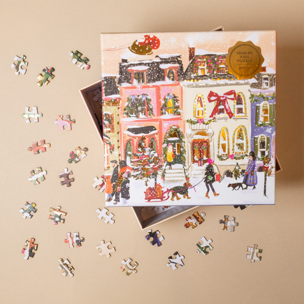 snowfall-on-main-street-1000-piece-puzzle Jigsaw puzzle box with a festive scene and scattered pieces on a beige background