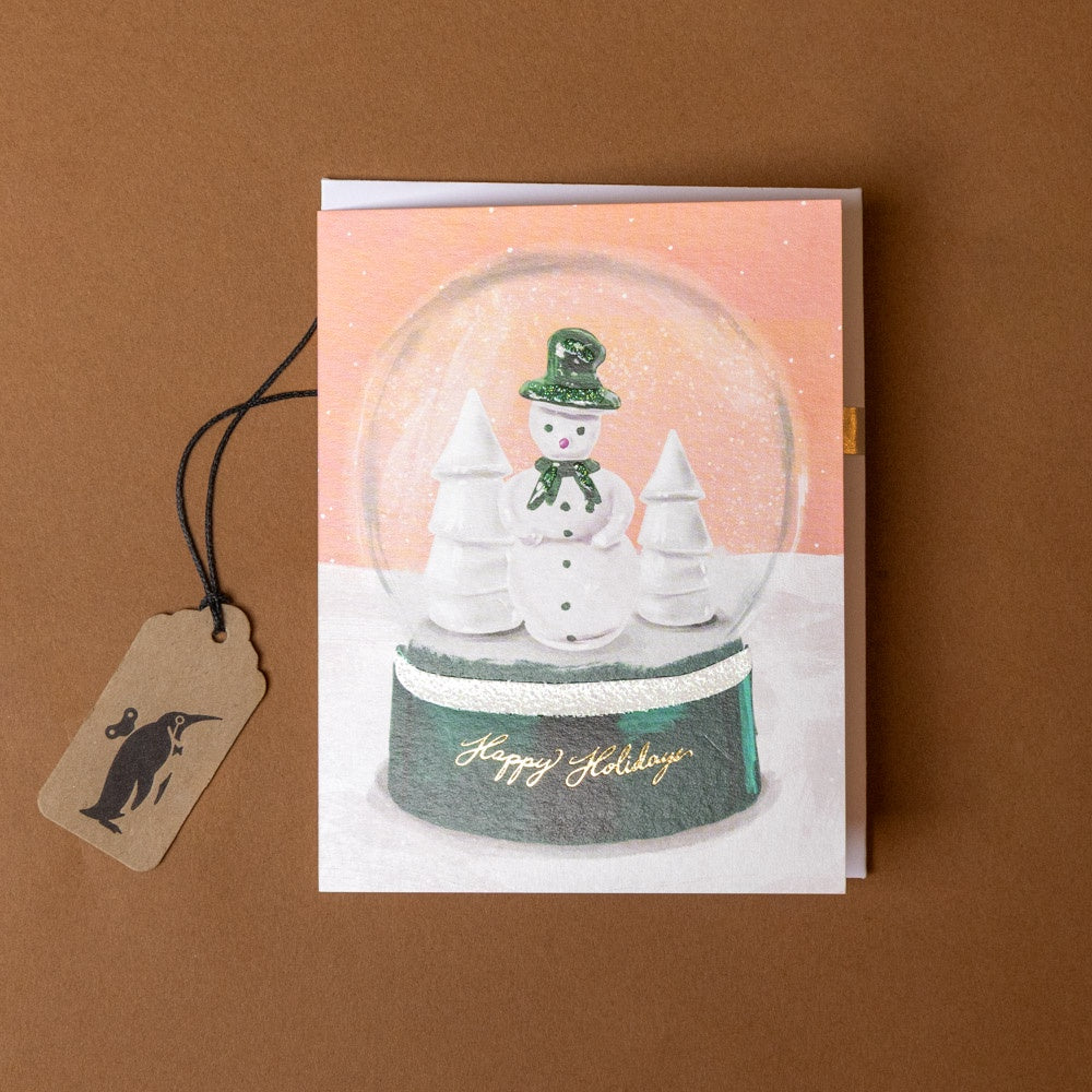 snowglobe-holidays-greeting-card with a snow globe design featuring a snowman and trees, on a brown background.